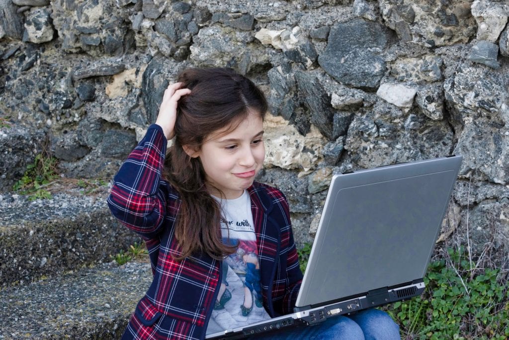 little girl sitting outside on stone steps scratching her head while looking at a laptop in a confusing way